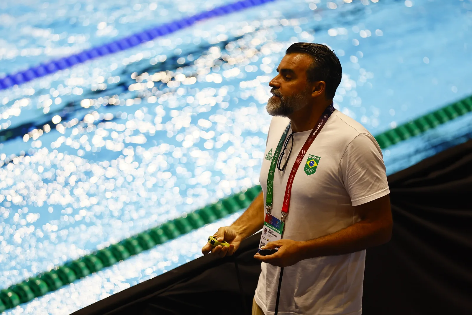 Un entrenador de natación al borde de la piscina con un cronómetro en cada mano, observando el agua.