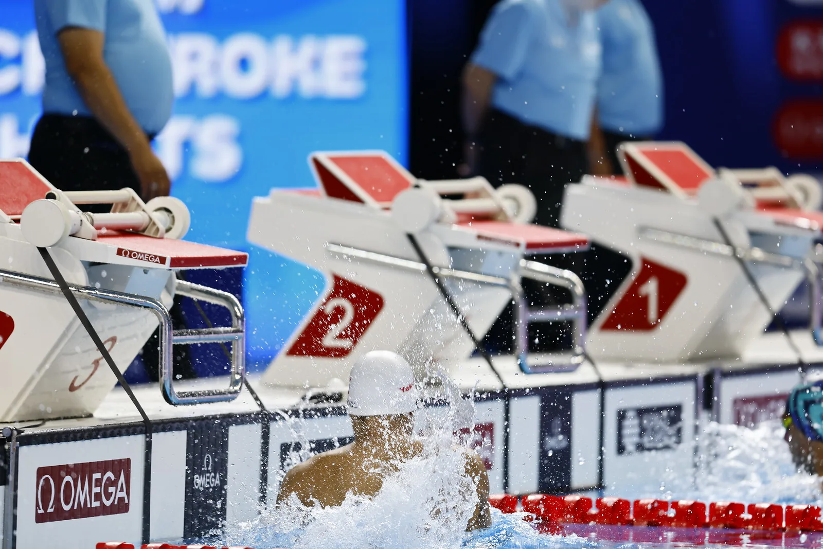 A swimmer touching the pool wall with a large splash, timing equipment visible behind.